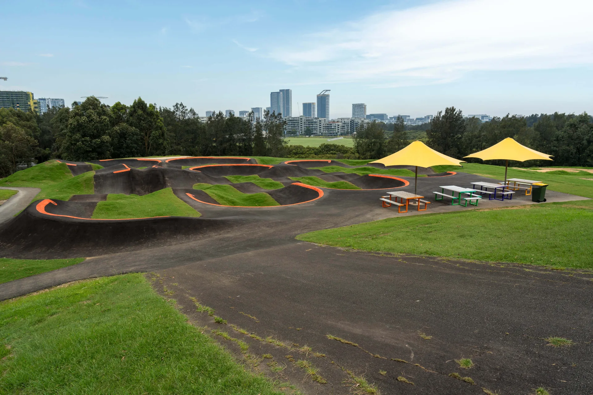 Asphalt pump track with orange-lined berms, featuring shaded picnic tables, yellow umbrellas, and grassy banks with a city skyline view.