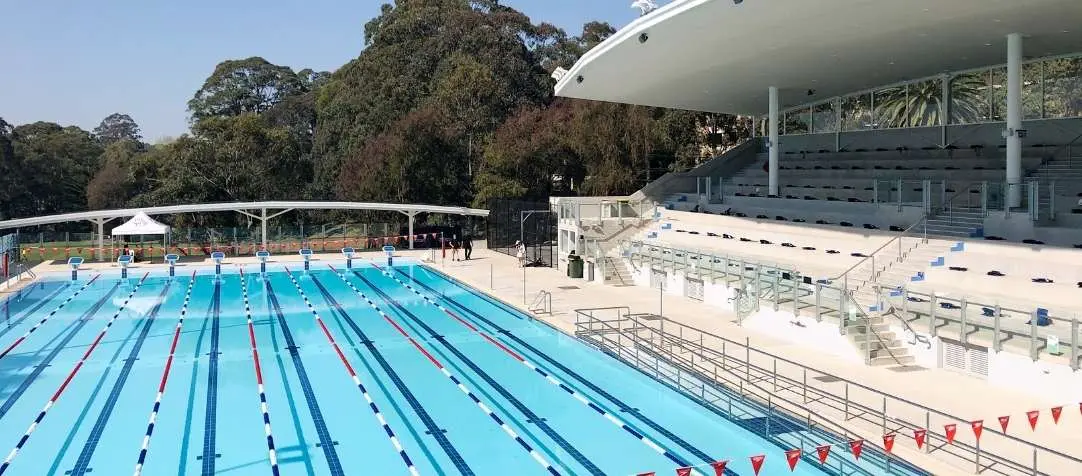 Large outdoor swimming complex with 8-lane competition pool featuring lane ropes, modern grandstand seating with white canopy roof, and surrounding parkland with mature trees.
