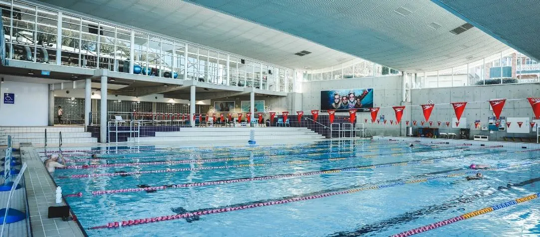 Indoor aquatic center with multiple lap lanes marked by colorful lane ropes, stadium seating with red banners, large windows providing natural light, and swimmers actively using the pool.