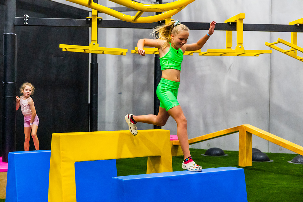 A child balancing across foam blocks beneath an overhead ninja rig on the X-Park course at BOUNCE Cromer.