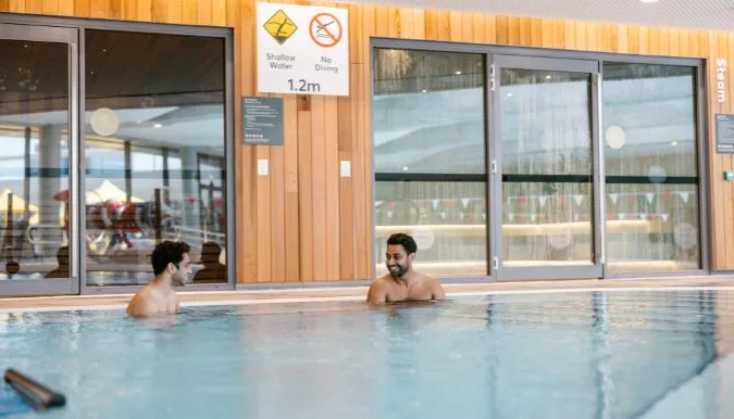 Two people relaxing in a warm indoor pool at Parramatta Aquatic Centre, with large windows and wooden exterior walls creating a modern, welcoming atmosphere.
