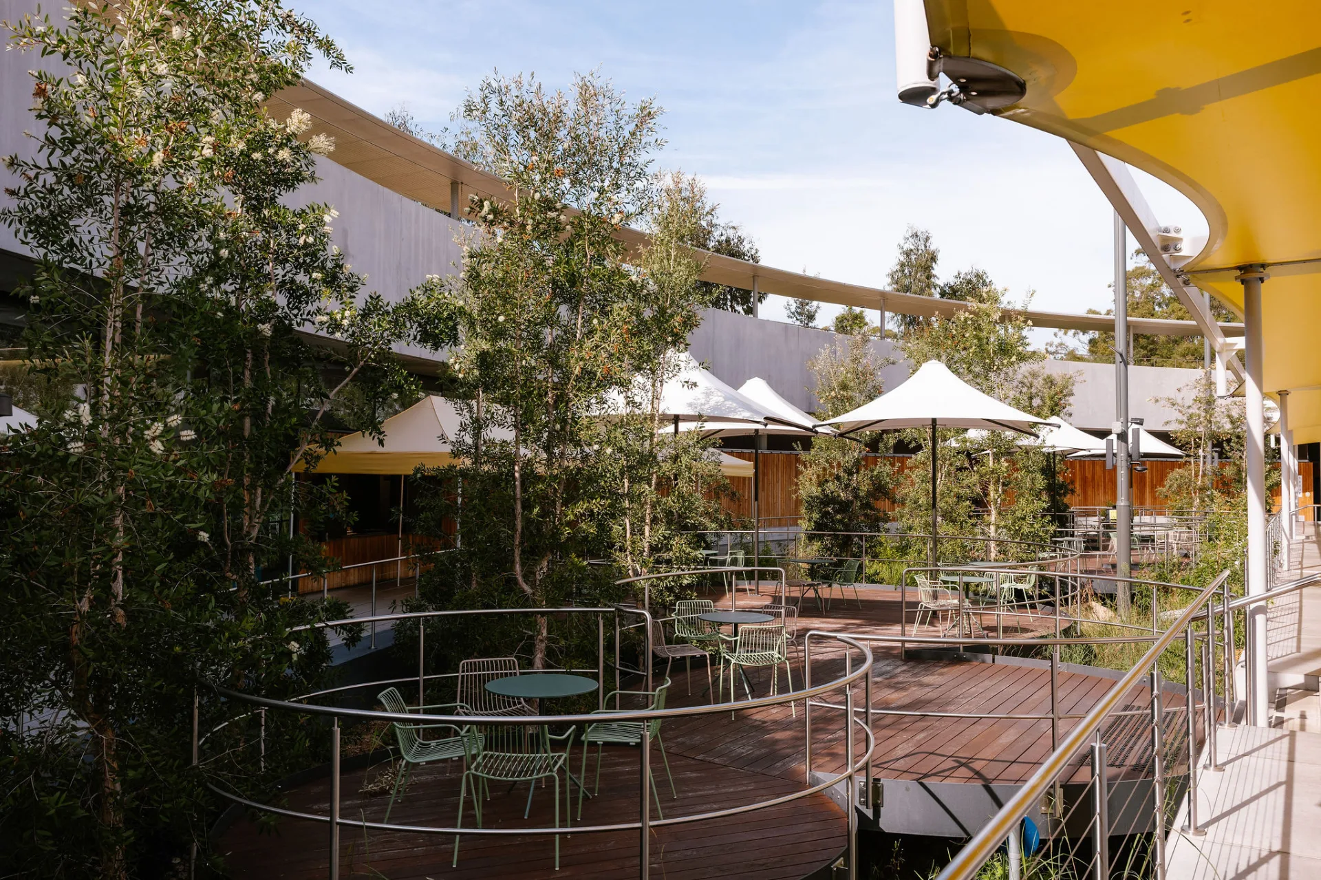 Multi-level wooden deck dining area with green wire chairs, white umbrellas, and metal railings surrounded by trees at aquatic center