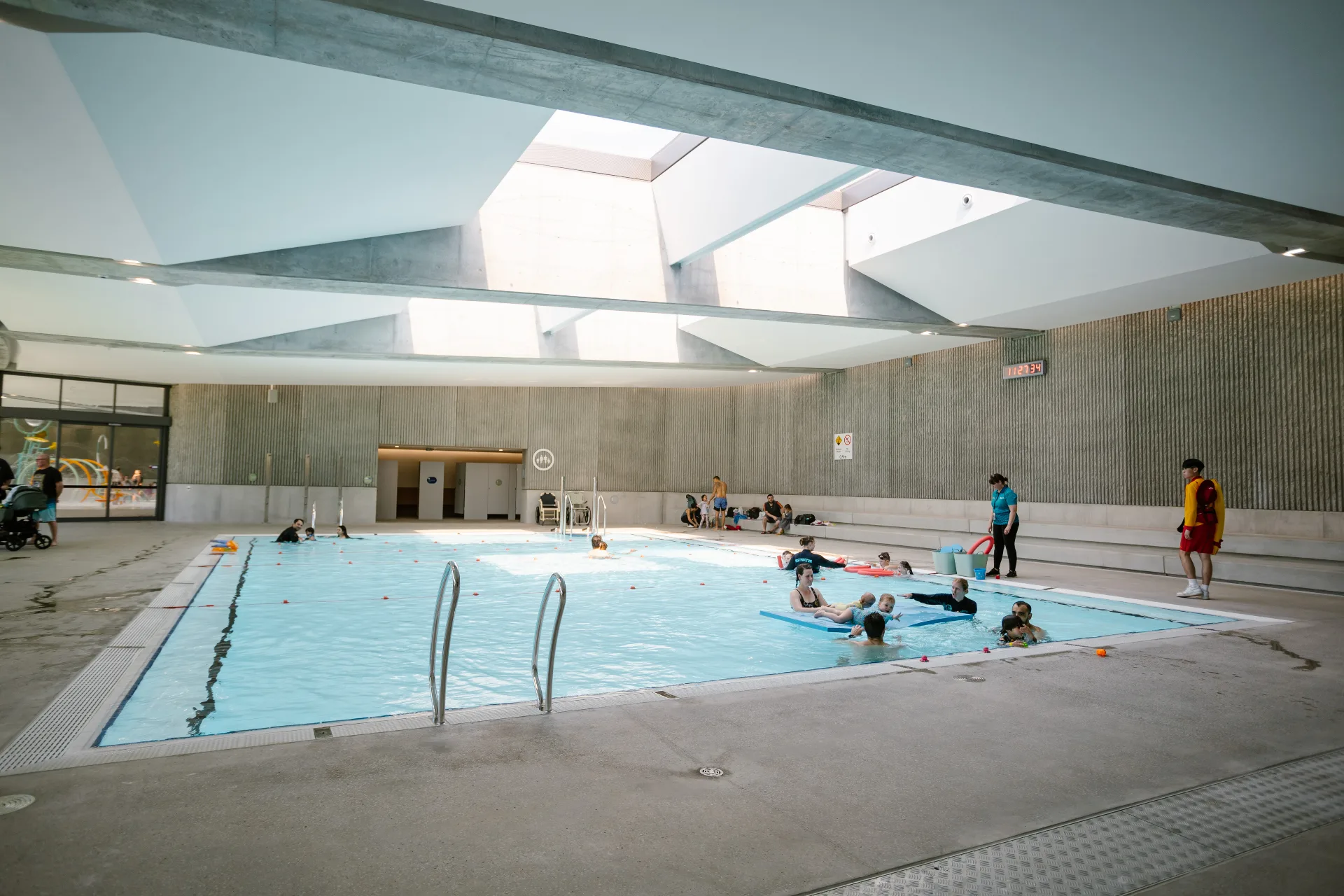 Indoor swimming pool with children's swim class in progress, featuring modern concrete architecture with glass ceiling panels and poolside instructors supervising activities.
