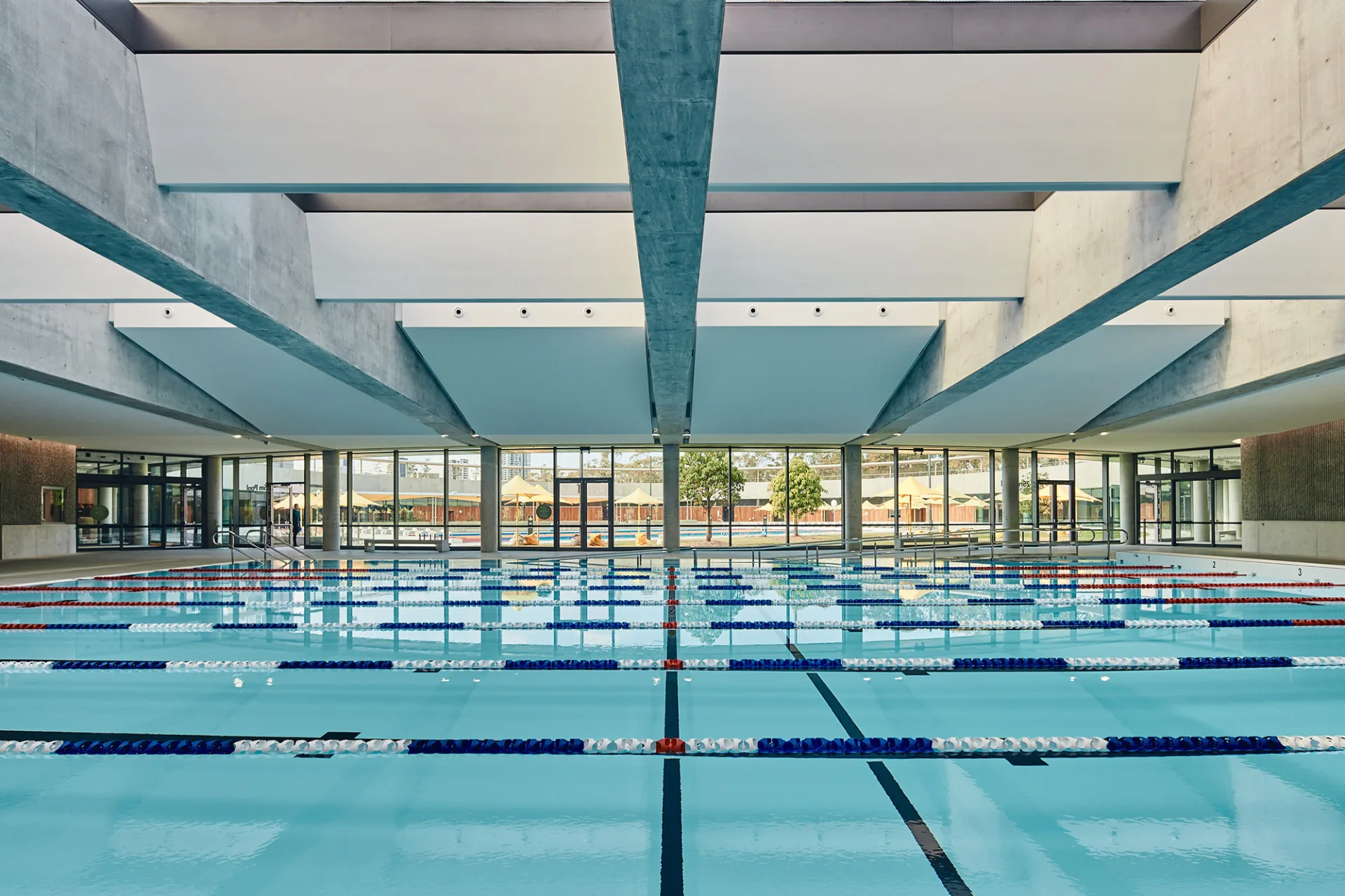 Indoor swimming pool with multiple lap lanes marked by blue and red lane ropes, large windows overlooking outdoor area, and modern concrete beam ceiling structure