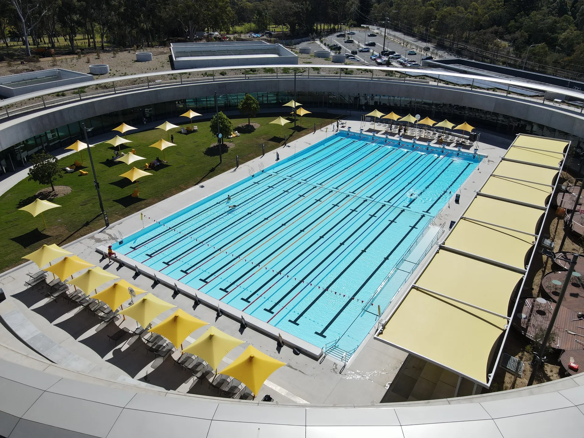 Modern outdoor aquatic center with Olympic-size swimming pool featuring multiple lanes, surrounded by yellow shade umbrellas, grassy areas, and contemporary curved architecture with viewing decks above.