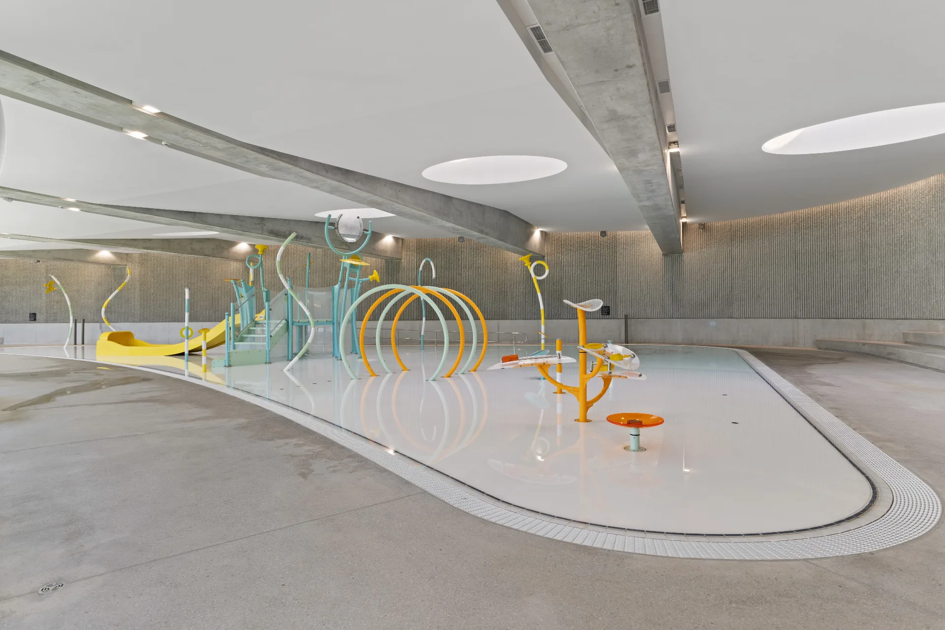 Modern indoor children's pool with colorful water play features including slides, spray fountains, and climbing structures in a bright aquatic center with curved concrete ceiling