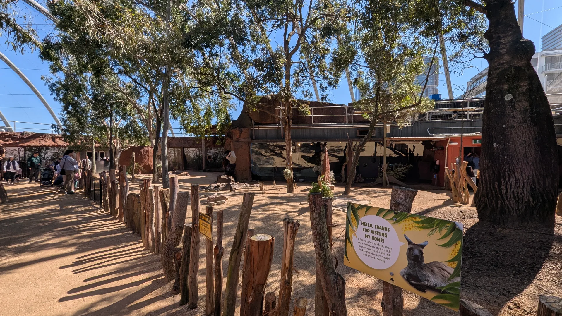 Outdoor zoo exhibit with wooden fence barriers, sandy pathways, and large shade trees. Visitors can observe animals in naturalistic enclosures with covered viewing areas.