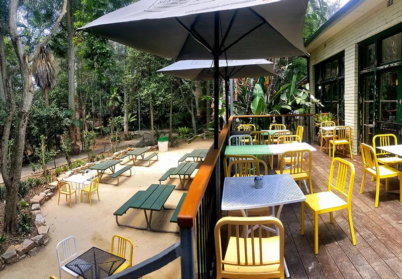 Outdoor cafe seating area with yellow chairs and tables on wooden deck, large umbrellas for shade, and children's playground equipment visible in background surrounded by lush trees.