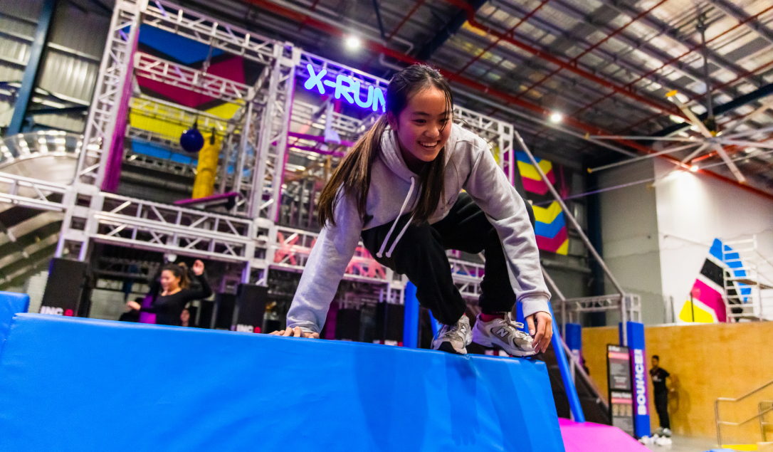 A child climbing over a padded obstacle on the X-Run ninja course with the X-Park course in the background at BOUNCE Homebush.