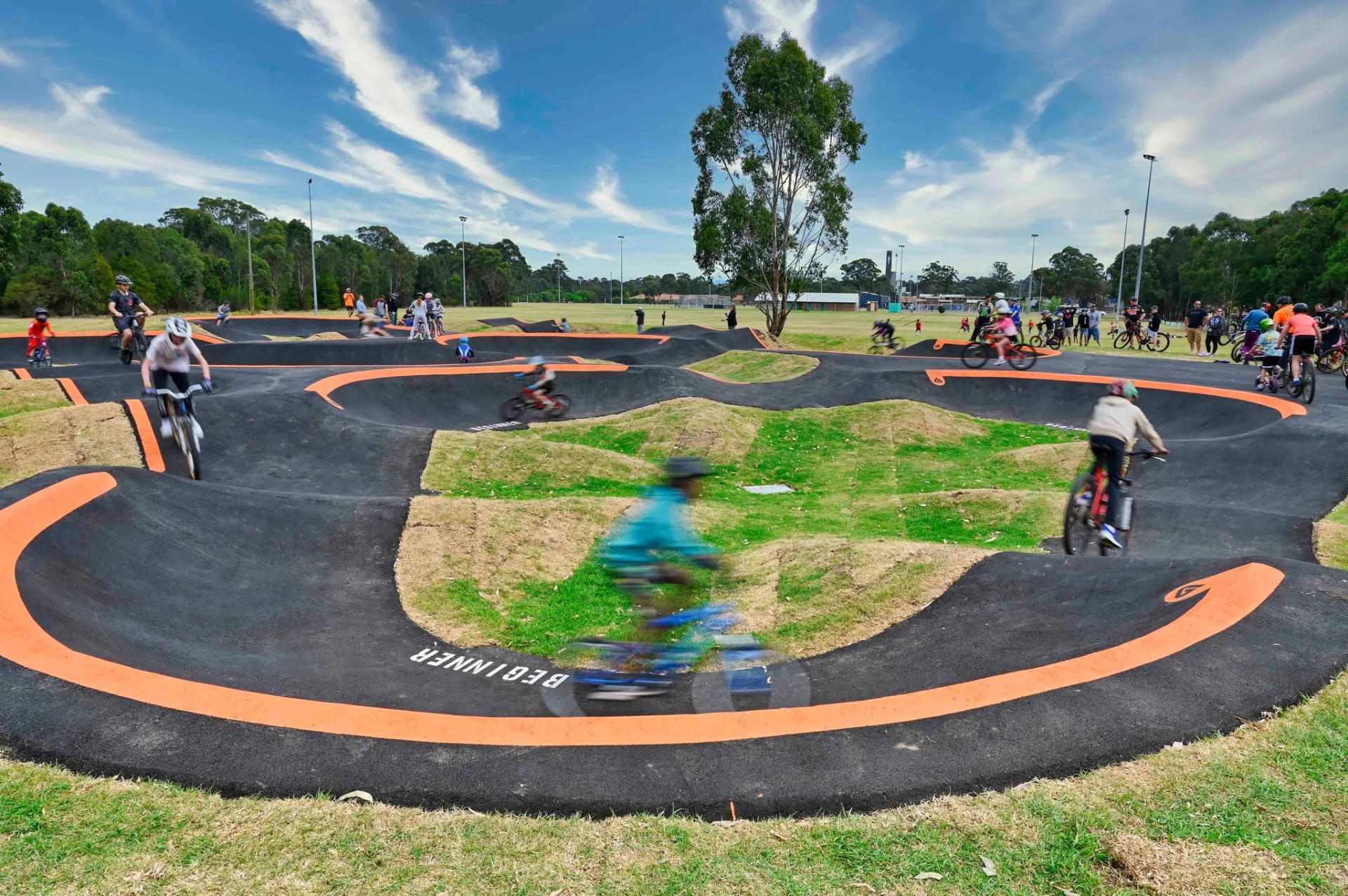 Curved asphalt pump track with orange borders where cyclists ride around banked turns and over small hills, surrounded by grass and trees under a blue sky.