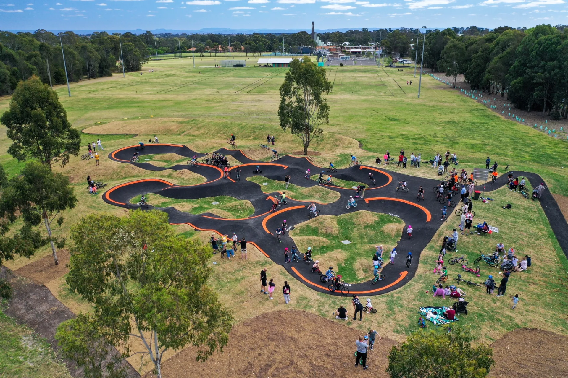 Aerial view of a curved black asphalt pump track with orange lane markings, surrounded by green parkland with families and cyclists gathered around watching riders navigate the winding course.