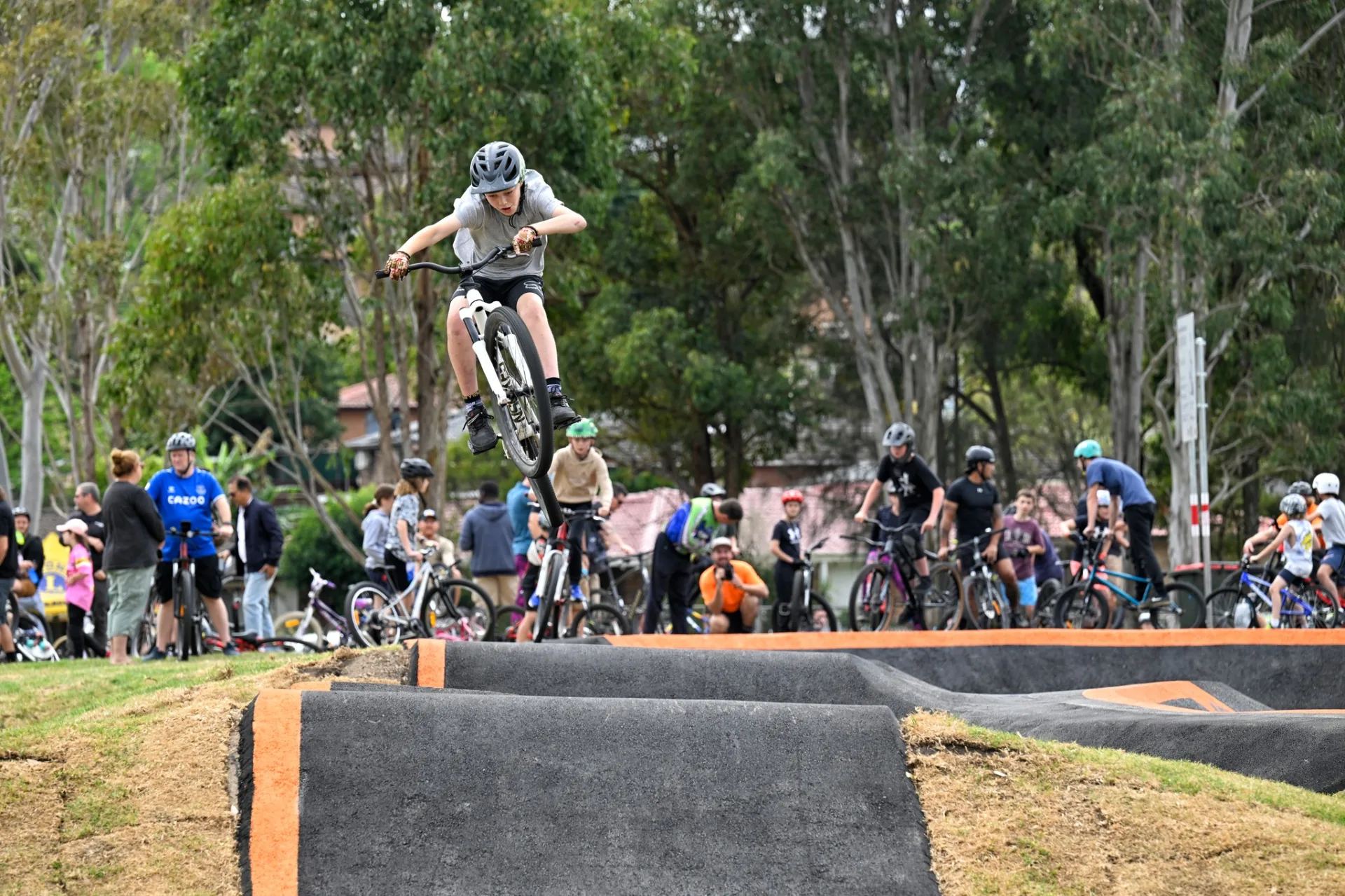 Young cyclist airborne on pump track jumps with crowd of families and bikes watching from grassy area surrounded by trees