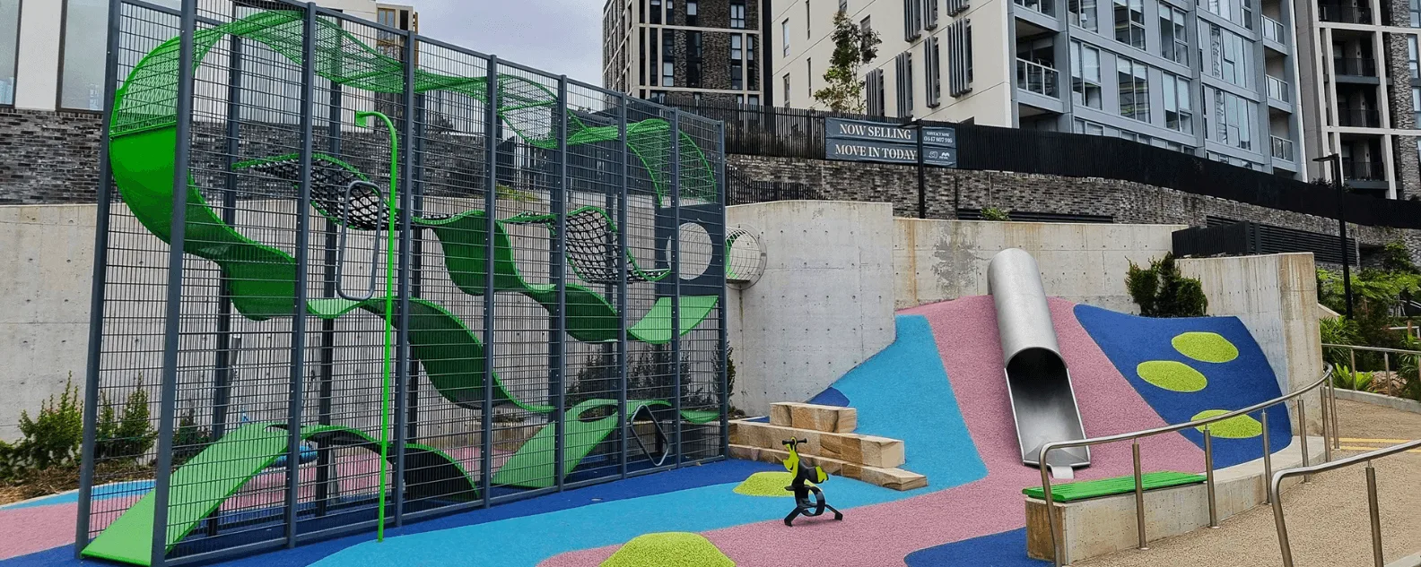 Colorful modern playground with tall metal slide, climbing equipment behind safety fencing, and vibrant blue and pink rubber surfacing with green circular patterns.