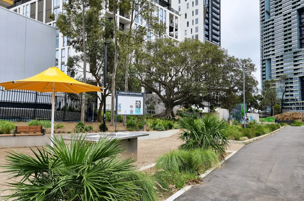 Modern urban park with wide paved pathways, native landscaping, yellow shade umbrella over seating area, and mature trees surrounded by high-rise apartments.