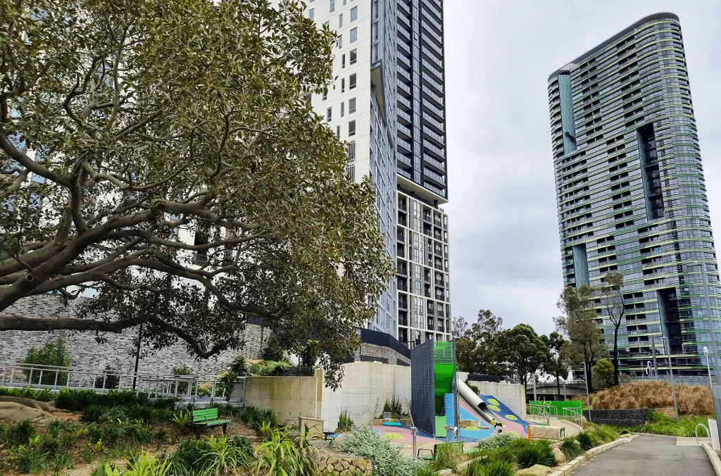 Colorful playground with slides and climbing equipment nestled between modern high-rise apartments, featuring mature shade trees and landscaped gardens with paved pathways.