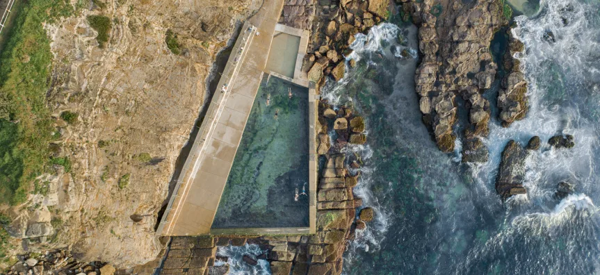 Aerial view of the Avalon ocean rock pool, a rectangular swimming pool set into rocky shoreline with swimmers inside and ocean waves breaking against the rocks alongside.