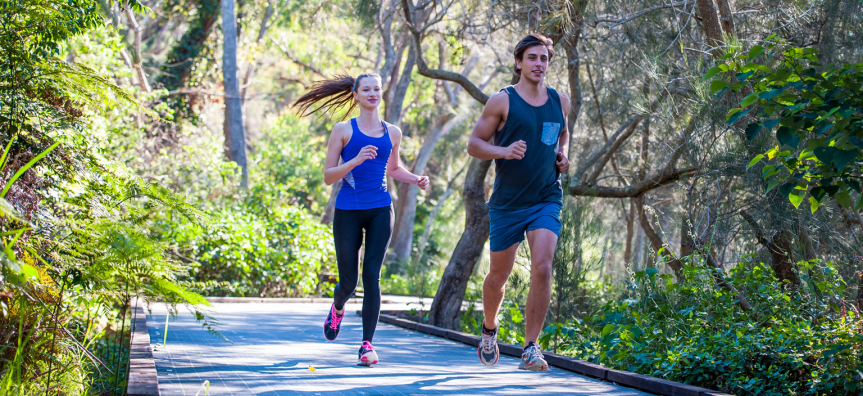 Two people running along a paved shared path lined with ferns and bushland on the Narrabeen Lagoon Trail.