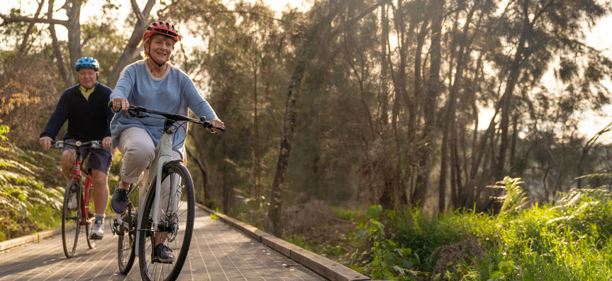 Two cyclists wearing helmets riding along the boardwalk section of the Narrabeen Lagoon Trail through bushland at sunset.