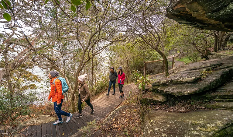 Family walks along wooden boardwalk through bushland with large sandstone rock formations, featuring handrails and natural canopy shade.