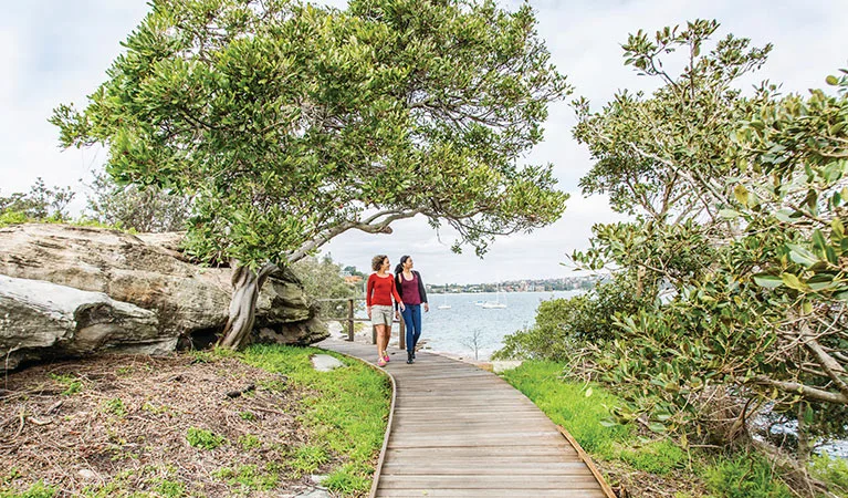 Two people walk along a wooden boardwalk beside Sydney Harbour, surrounded by native trees and sandstone rocks with water views in the background.