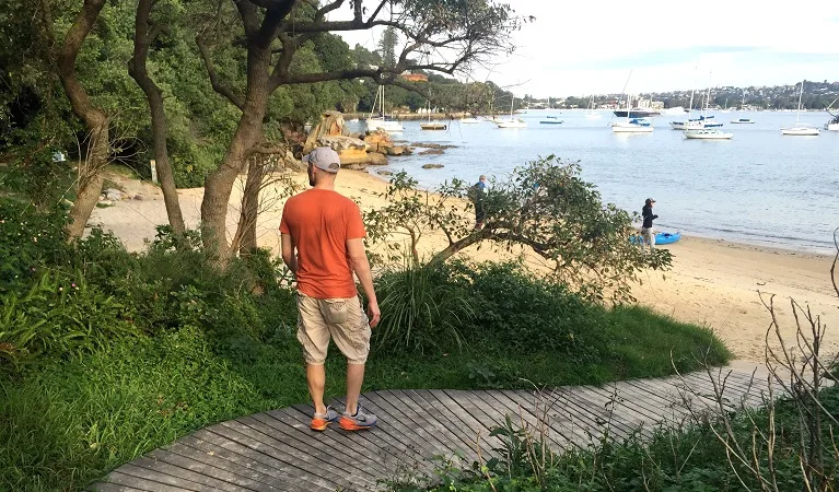 Person in orange shirt standing on wooden boardwalk overlooking calm bay with moored boats, sandy beach, and coastal vegetation providing natural shade along the waterfront track.