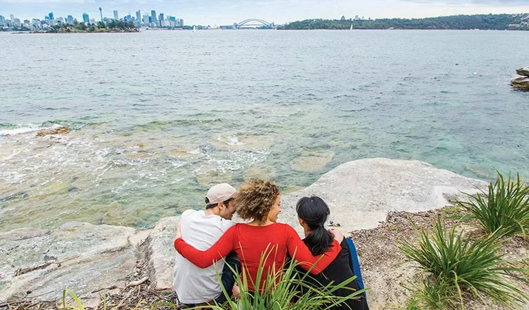 Three people sit together on sandstone rocks overlooking clear harbor waters with Sydney city skyline in background, enjoying a family-friendly waterfront spot with natural seating areas.