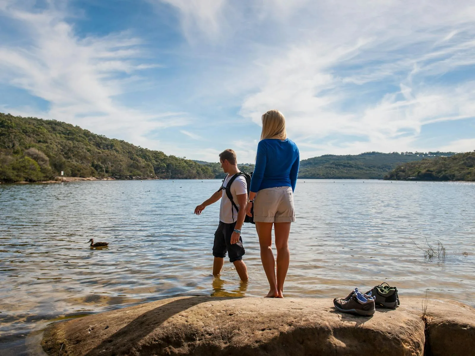 Woman and child wading in calm lake waters at Manly Dam, surrounded by forested hills with hiking shoes on rocky shore nearby