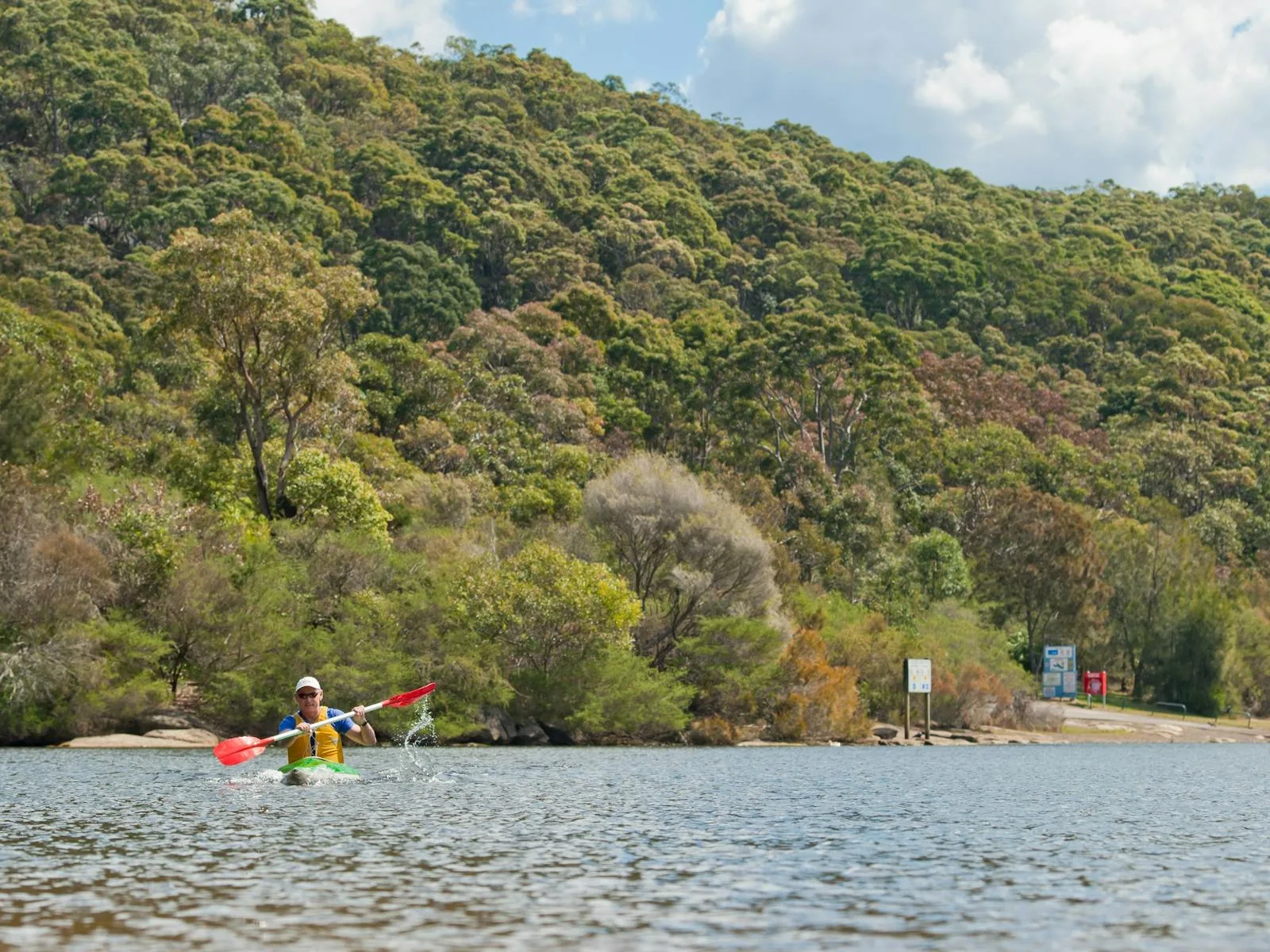 Kayaker paddles across calm lake waters with forested hills and information signs visible on the wooded shoreline at Manly Dam recreation area.