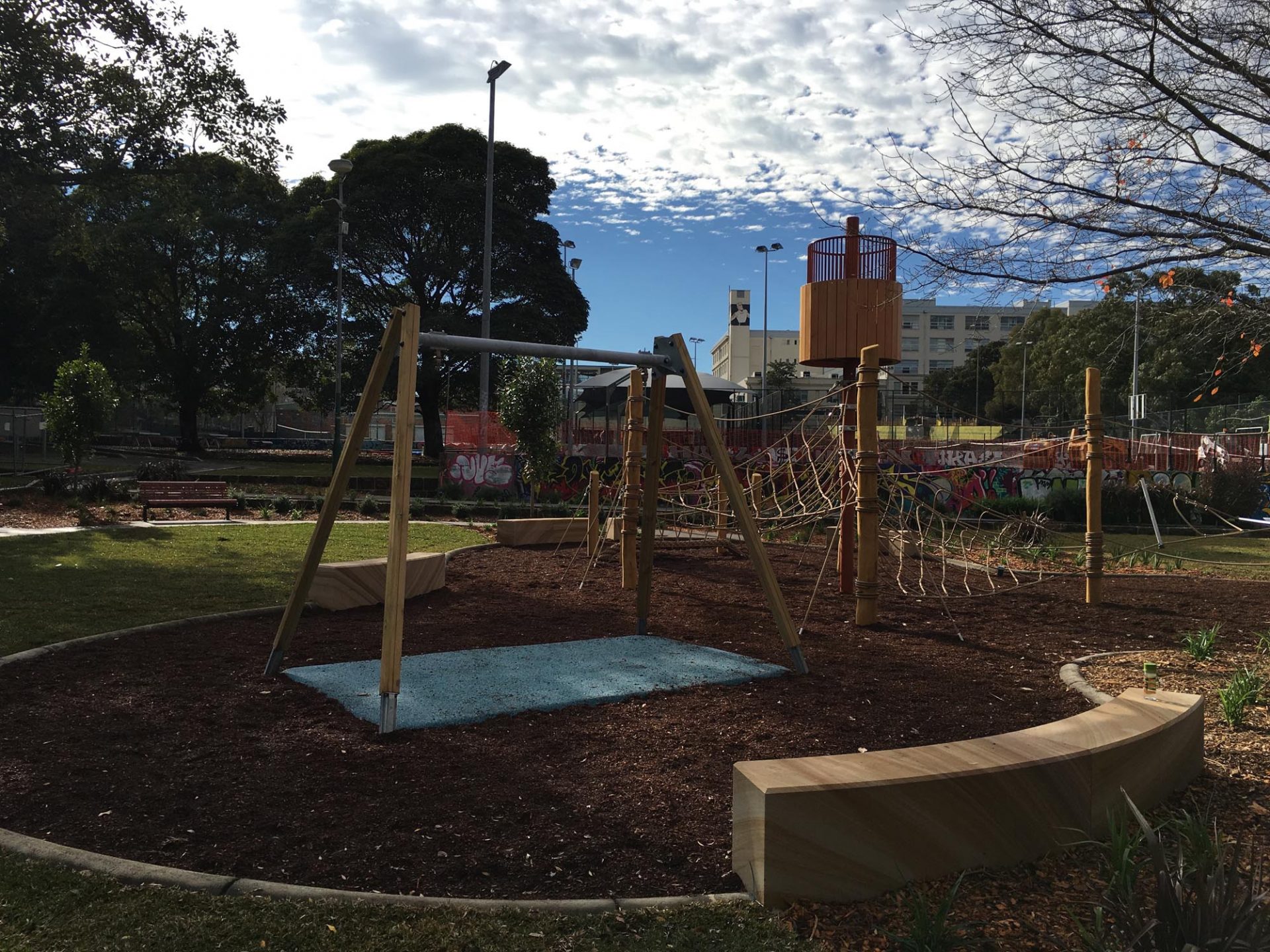 Nature-based playground with a timber swing frame over a blue soft-fall mat, a rope climbing net and an elevated timber lookout tower, edged by curved timber seating and bark mulch.