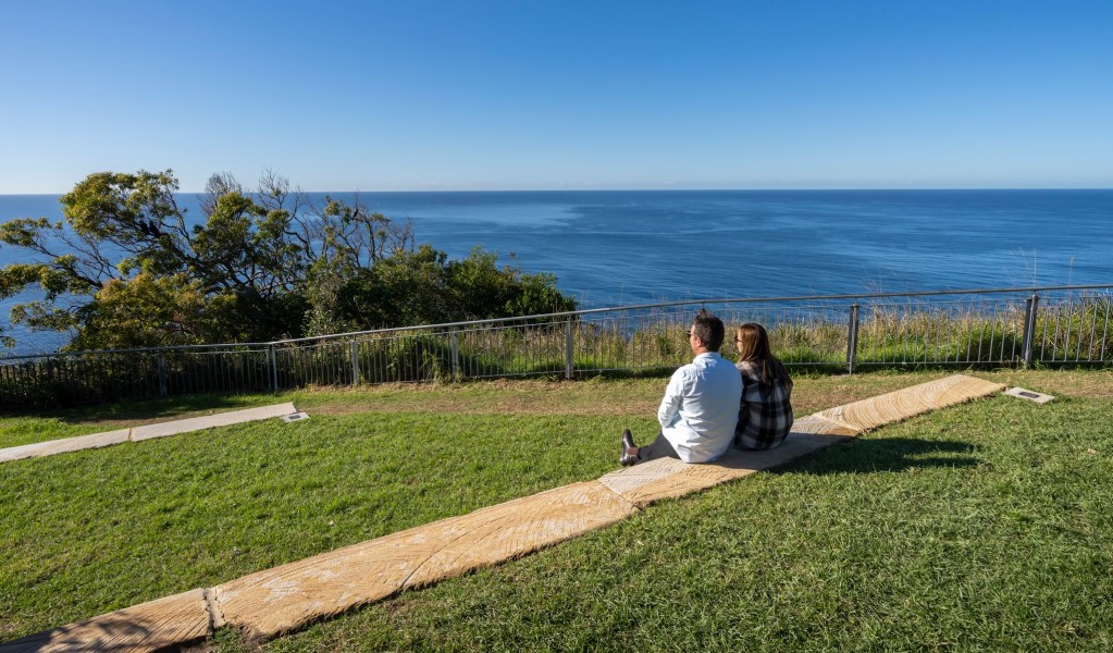 A couple sitting on the grass looking out to the ocean from Crackneck Point Lookout