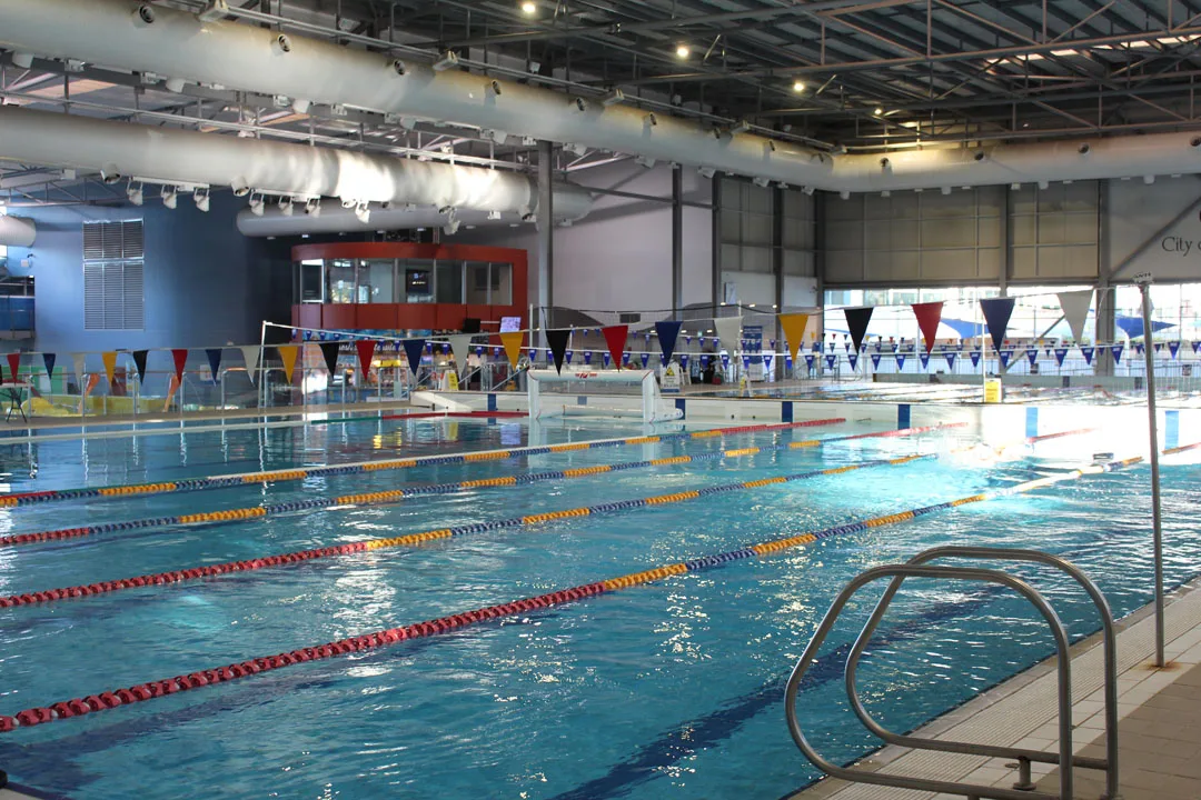 Indoor swimming pool with multiple lap lanes marked by colorful rope dividers, triangular pennant flags overhead, and pool ladder visible in foreground