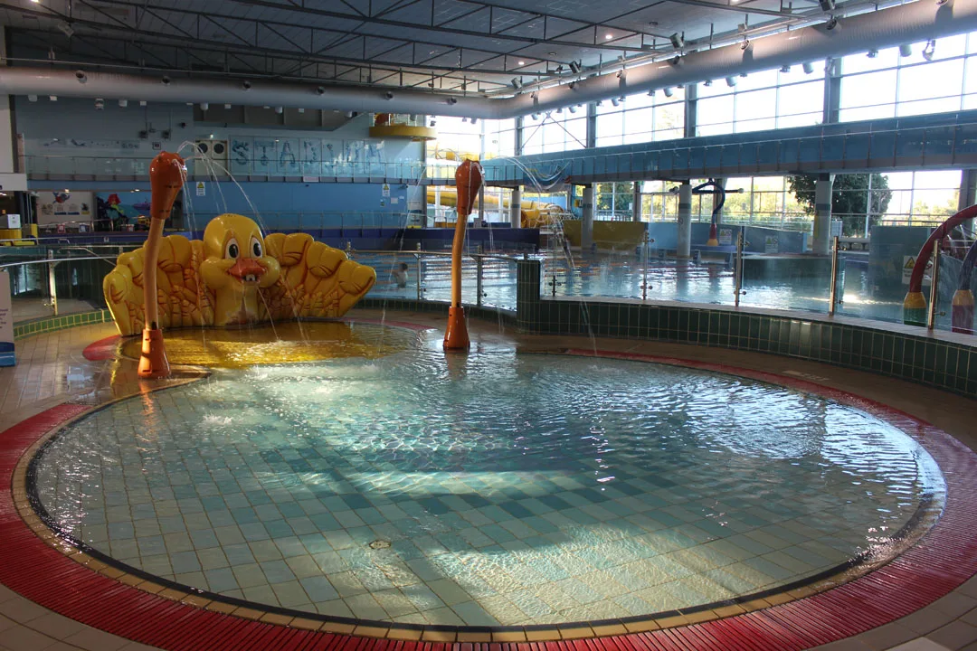 Indoor children's pool area with shallow circular wading pool, yellow duck-shaped water feature, and orange water play equipment under bright skylights at aquatic center