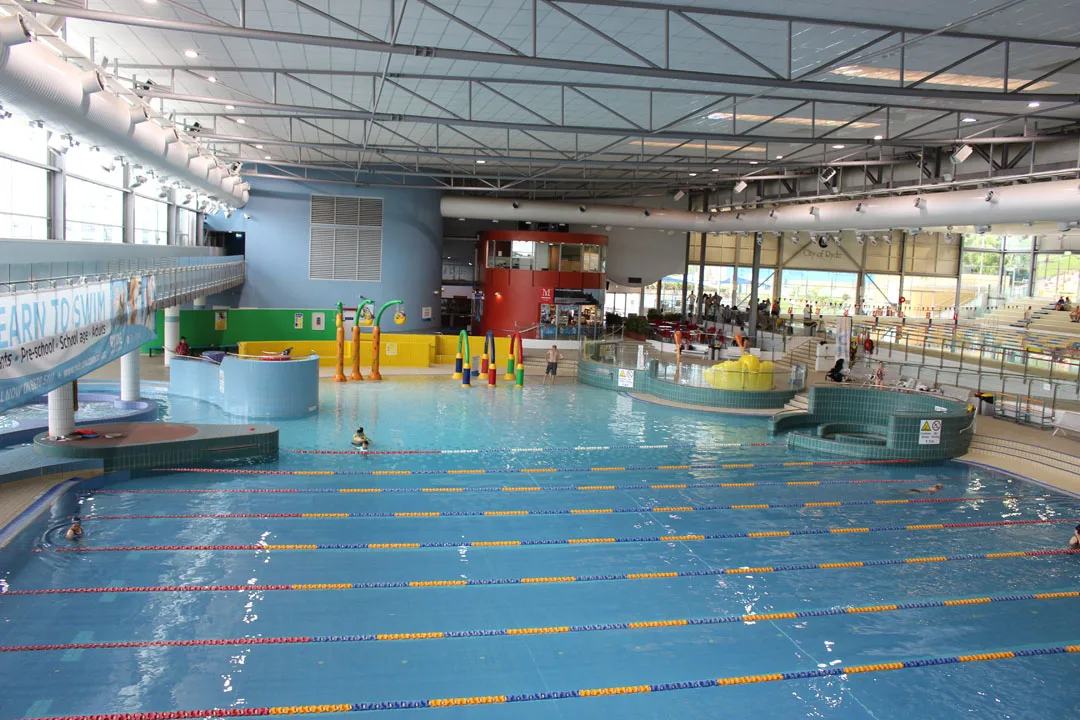 Indoor aquatic center with lap pool lanes, colorful children's play area featuring water features and slides, plus tiered seating for families under a glass roof structure.