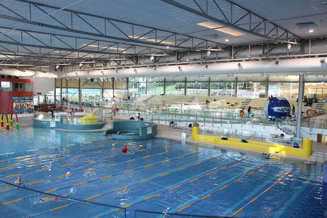 Large indoor aquatic center with lap pool featuring lane ropes, children's play area with colorful water features, stadium seating, and floor-to-ceiling windows providing natural light.