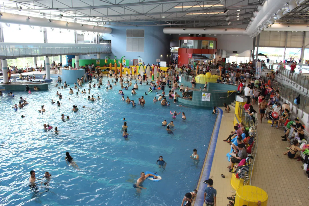 Busy indoor aquatic center with large swimming pool filled with families, colorful water play equipment, and spectator seating area along the poolside