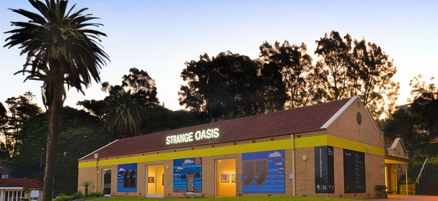 Exterior of Manly Art Gallery & Museum at dusk on West Esplanade, with a large exhibition banner reading Strange Oasis across the building.