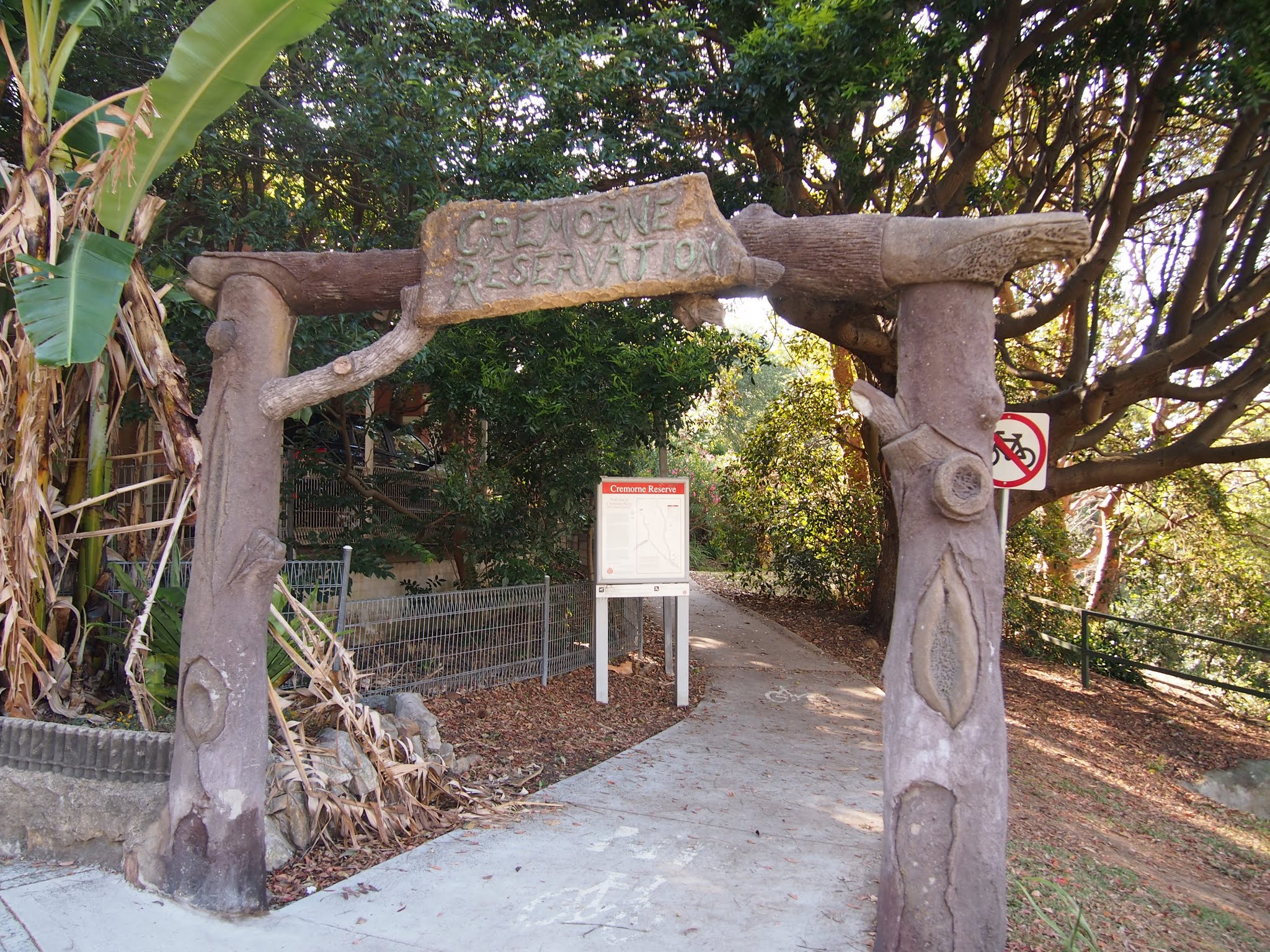 Carved timber archway reading "Cremorne Reservation" over a paved path leading into the bushland of the foreshore reserve.