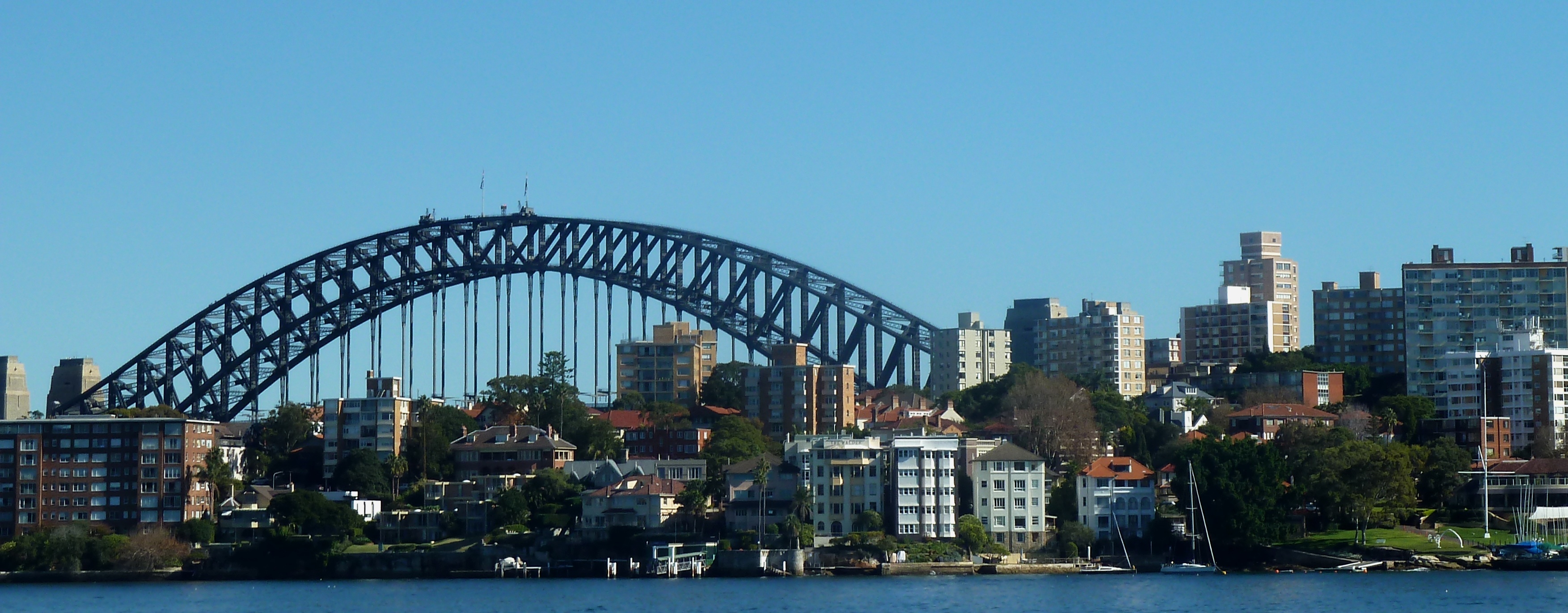 View across Sydney Harbour from Cremorne Point showing the Sydney Harbour Bridge arch behind a waterfront row of houses and apartment blocks.