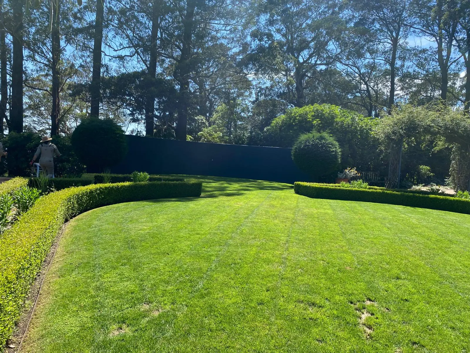 Well-maintained formal garden with manicured circular lawn surrounded by neat hedging, mature trees providing shade, and a groundskeeper visible maintaining the space.
