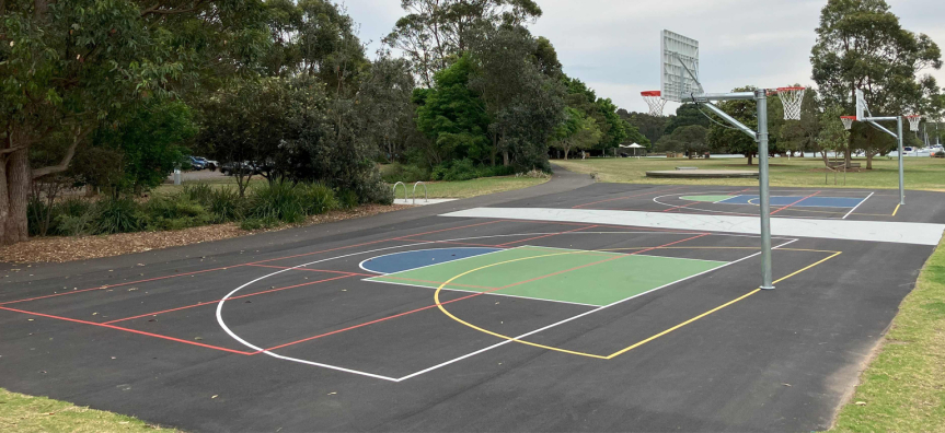 Outdoor multi-use sports court with basketball hoops and coloured line markings, bordered by parkland and trees.