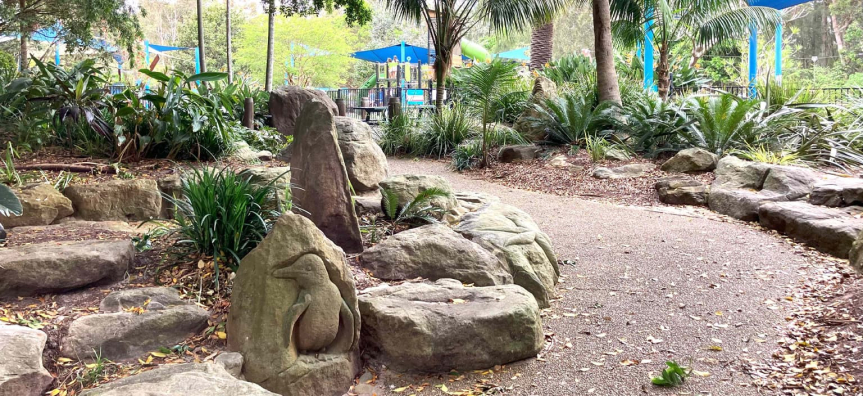 Landscaped garden path lined with sandstone rocks carved into sea creatures, with the playground's blue shade sails behind.