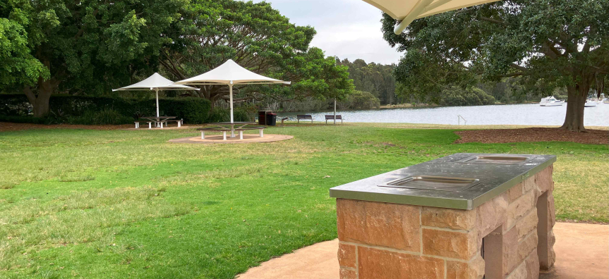 Sandstone barbecue in the foreground with shaded picnic tables on the lawn and a boat on the calm Pittwater bay behind.