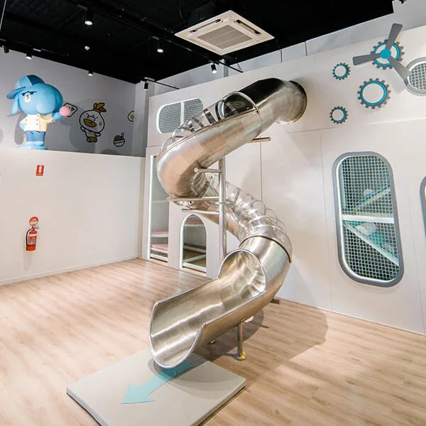 Indoor play area with metallic spiral tube slide connecting upper and lower levels, featuring spaceship-themed white walls with portholes and colorful cartoon characters.