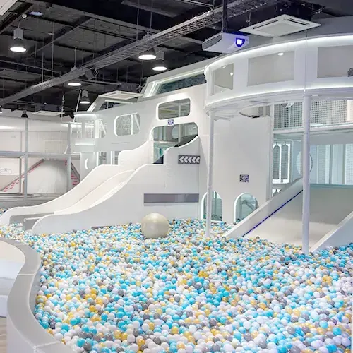 White futuristic play structure with slides and climbing areas surrounded by a large colorful ball pit in an indoor children's play center with modern lighting