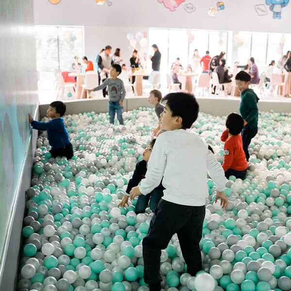 Children playing in a large ball pit filled with green, white, and gray plastic balls at an indoor play center with parents watching from seating areas in the background.