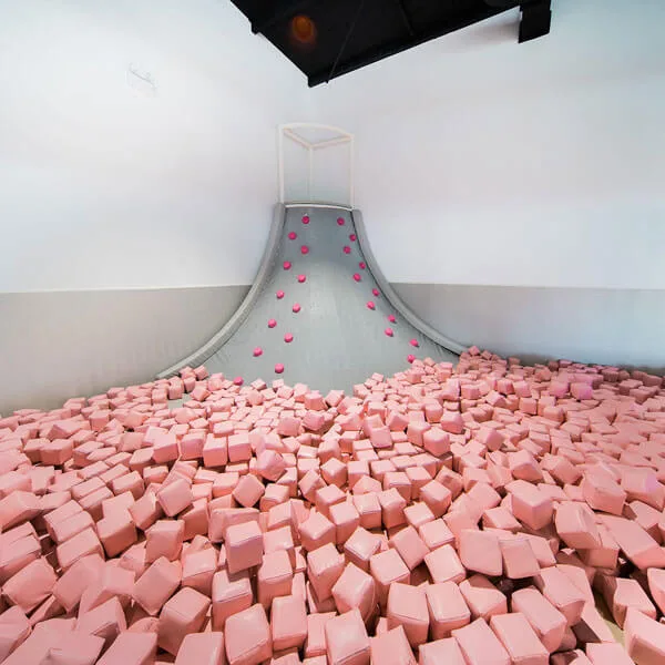 Indoor play area with large white slide leading into pit filled with thousands of pink foam blocks, with pink spheres decorating the slide walls