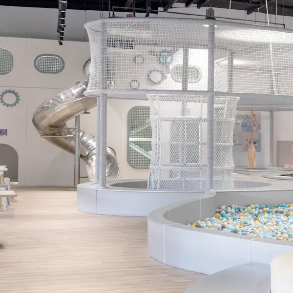 Modern white indoor play structure with multi-level climbing area, spiral slide, and colorful ball pit in bright children's play center