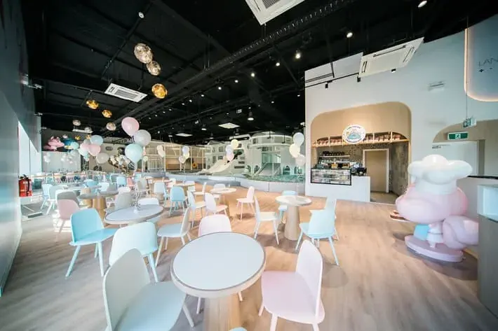 Modern indoor play center with pastel-colored seating areas, colorful balloon decorations, multi-level play structures with slides, and a cafe counter in an open-concept space with high ceilings.