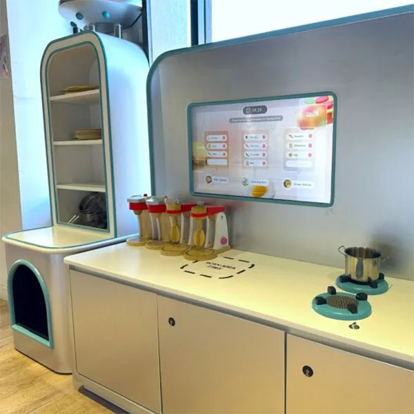 Modern white play kitchen station with digital touchscreen menu, storage compartments, colorful cups, and interactive cooking accessories for children's pretend play.