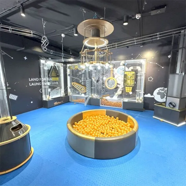 Interactive space-themed play area with blue padded flooring, circular ball pit filled with orange balls, and transparent climbing structure with space station elements overhead